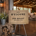 a wooden sign with a welcoming message for a pig roast celebration, placed in front of a rustic wooden structure with a lantern and flowers.