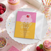 A birthday party invitation for a girl named Amy, who is turning three, with details about the party on a white card with a pink and yellow background. The invitation is placed on a white plate, surrounded by various desserts, such as cupcakes, raspberries, and sprinkles.