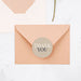 a peach-colored envelope with a round "Thank You" sticker on it, placed on a white surface.