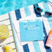 a blue invitation card for a "SPLISH SPLASH" party, placed on a towel next to a pair of sunglasses and a glass of lemonade.