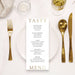 a menu with gold lettering, placed on a table with a fork, knife, and glass of wine.