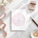 a wedding invitation card with a pink and gold geometric design, surrounded by wedding-related items such as a ring box, a pen, and some gold confetti.