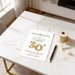 A white wedding invitation card with gold numbers sits on a white marble table, accompanied by a pen and a roll of tape.