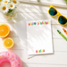 A white table with various items on it, including a note with a personalized message, sunglasses, a pink donut, a glass of water, and a vase of flowers.