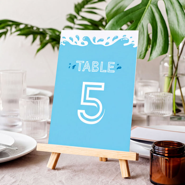 A blue table number sign displaying the number 5, placed on a wooden easel on a table with various glassware and a plant in the background.