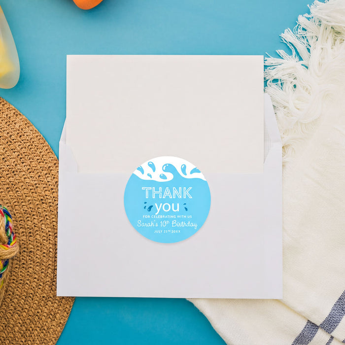 a white envelope with a blue and white "Thank you" sticker on it, placed on a blue surface with a straw hat and a white towel nearby.