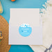 a white envelope with a blue and white "Thank you" sticker on it, placed on a blue surface with a straw hat and a white towel nearby.