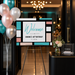 a colorful sign welcoming guests to a birthday celebration for someone named Simone. The sign is displayed on an easel, with balloons and a vase of flowers nearby.