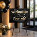a black sign with gold text that reads "Welcome to Olivia's Fabulous Fourth Birthday Celebration" and "DECEMBER 2ND 20XX". The sign is placed on a stand, and there are black and gold balloons in the background.