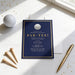 a blue invitation card with a golf ball design, placed on a white surface alongside a golf ball, a pen, and some scattered gold glitter.