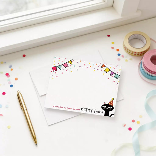 a birthday card with a cat wearing a party hat, surrounded by confetti and a pen on a white surface.