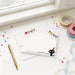 a birthday card with a cat wearing a party hat, surrounded by confetti and a pen on a white surface.
