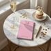 a pink notebook with the name "Juliette Wills" on it, placed on a marble table. Next to the notebook is a cupcake with a candle, and a glass of champagne. There are also gold confetti scattered around the table.