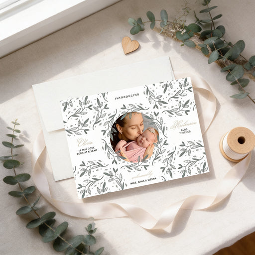 a white card with a floral design, a photo of a woman holding a baby, and some greenery and a ribbon.