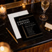 a black invitation card for an event called "EMERSON LAWYERS' ANNUAL DINNER" with details about the date, time, location, and other event information. The card is placed on a marble surface, with a glass of wine, a pen, and a business card nearby.