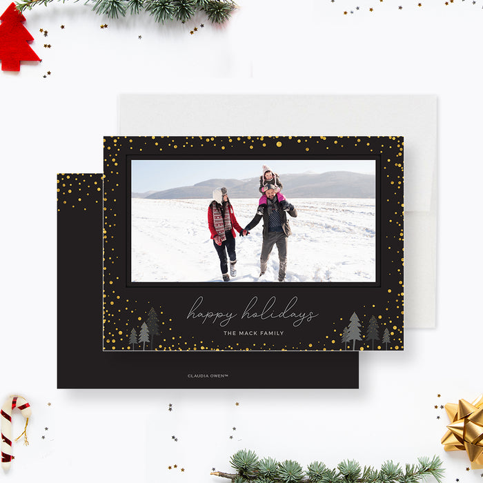 Modern black holiday photo card featuring gold confetti accents, a wide photo frame, scripted Happy Holidays greeting, and small illustrated winter trees.