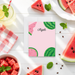 a pink card with a watermelon design, surrounded by various fruits and a drink on a white wooden surface.