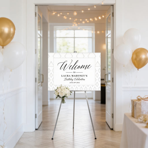 a white sign with gold lettering welcoming guests to Laura Mahoney's birthday celebration. The sign is displayed in a hallway with a floral arrangement on the floor in front of it. The hallway is decorated with gold balloons, and there is a table with a gift box on it.