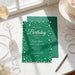 a birthday party invitation with a green background and pearls, placed on a table with a glass of wine, pearls, and a pen.