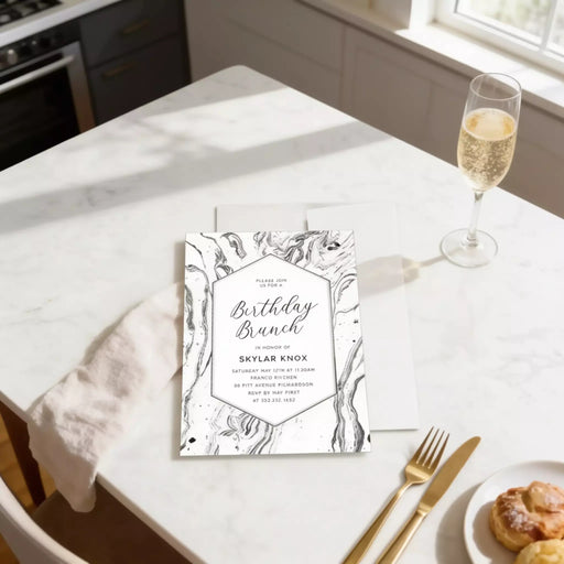 a birthday brunch invitation card for a celebration, placed on a white marble countertop next to a glass of champagne and a plate of pastries.