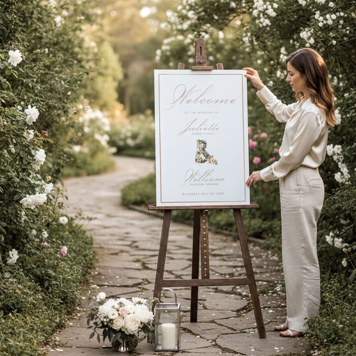 A woman is standing next to a wooden easel, placing a sign on it. The sign is white and reads "Welcome to the Wedding of Jacqueline and William". The woman is wearing a white blouse and beige pants. There are flowers and a lantern on the ground near the easel.