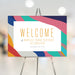 Colorful abstract birthday welcome sign with customizable name and date, featuring pink, teal, yellow, and blue, displayed on an easel.