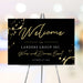 Black corporate wine dinner welcome sign with gold wine glass ring accents, script lettering, company name and event date