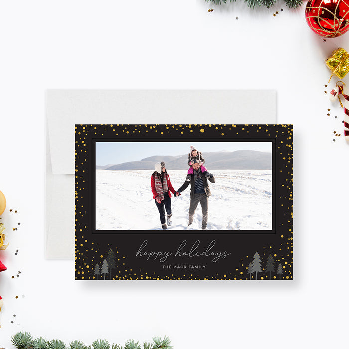 Modern black holiday photo card featuring gold confetti accents, a wide photo frame, scripted Happy Holidays greeting, and small illustrated winter trees.