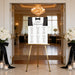 Personalized black and white black tie wedding seating chart sign displayed on a gold easel at an elegant reception entrance.