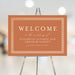 Burnt orange wedding welcome sign featuring an intricate border design with customizable names and date, displayed on an easel.