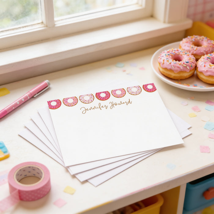 a desk with a stack of personalized note cards, a plate of pink frosted donuts, and a pink pen.