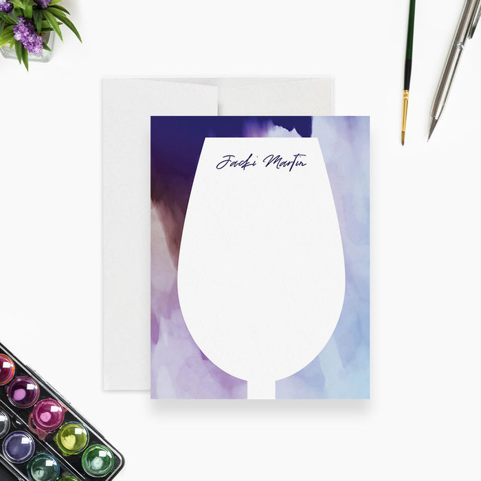 a blank card with a wine glass design, accompanied by a pen, a watercolor palette, and a small plant.