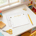 A white desk with a yellow pencil, a yellow stapler, and a yellow binder, along with a blank card and a small toy.