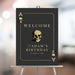 Black casino night birthday welcome sign styled as an ace playing card with gold skull illustrations, honoree’s name and event date
