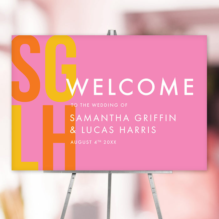 Modern pink wedding welcome sign with bold yellow and orange initials, customizable names, and date, displayed on an easel.