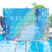 Personalized blue painterly pool party welcome sign with name and date, displayed on an easel at a poolside birthday event.