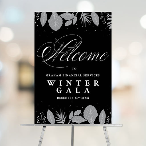 corporate winter gala welcome sign displayed on an easel with black background, gray botanical accents, elegant script lettering, and custom event details