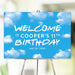 Personalized sky-themed birthday welcome sign with blue background and fluffy white clouds, custom name, age, and date, displayed on easel for party décor.