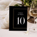 A black table number card with the number 10 displayed on it, placed on a table next to a glass of wine.