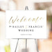 Minimalist wedding welcome sign with gold script and black text on white background, displayed on an easel at a wedding venue.