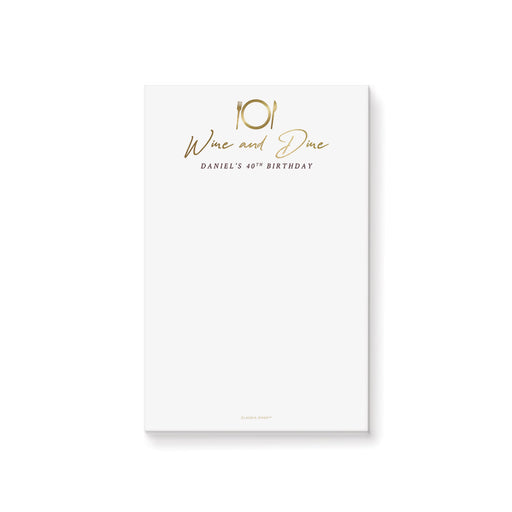 A personalized notepad with white background and gold text that reads 'Wine and Dine' and includes a name