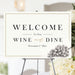 Wine and Dine welcome sign with grape illustration and customizable text, displayed on an easel for party or event decoration.