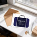 a desk with a computer monitor, a brown envelope, a blue suitcase with the text "We've MOVED" and a keychain, along with a pen and a gift box.
