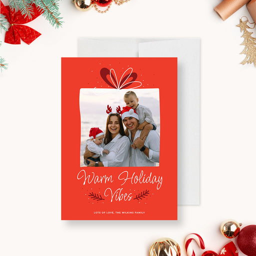 Family holiday photo card template featuring a red background, gift bow illustration, framed family photo and Warm Holiday Vibes greeting in a festive Christmas design