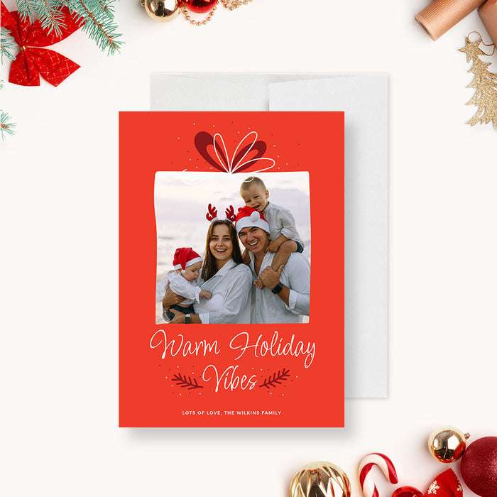 Family holiday photo card template featuring a red background, gift bow illustration, framed family photo and Warm Holiday Vibes greeting in a festive Christmas design