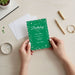 A person's hands holding a green birthday party invitation with white polka dots, surrounded by various office supplies and personal items on a desk.