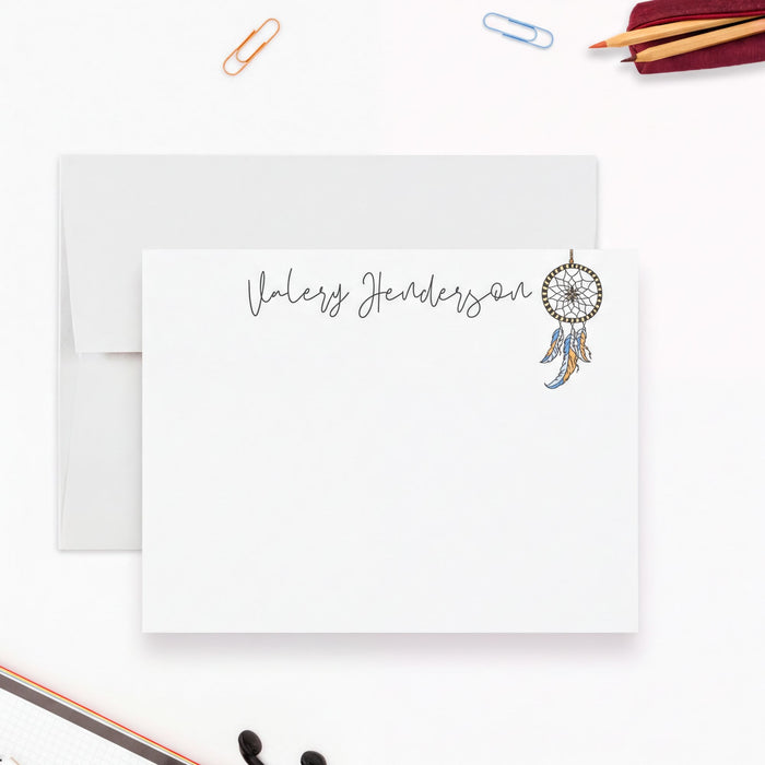 a blank, personalized note card with a name printed on it, along with a dreamcatcher illustration. The note card is placed on a white surface, surrounded by various office supplies such as a stapler, pencils, and a pair of scissors.