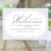 Floral garden party welcome sign on an easel featuring a lavender background with white flowers and elegant black script