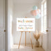 Baby shower welcome sign on an easel with balloons in a bright room.