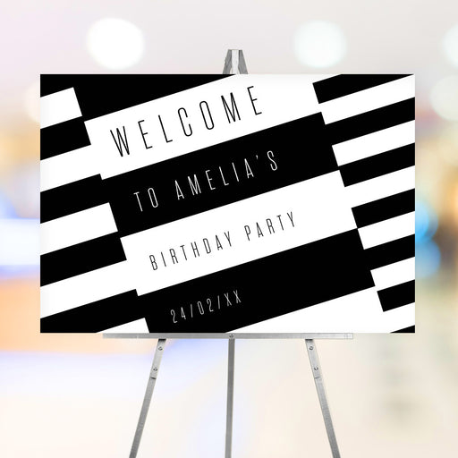 Modern black-and-white striped birthday welcome sign with minimalist typography displayed on an easel.